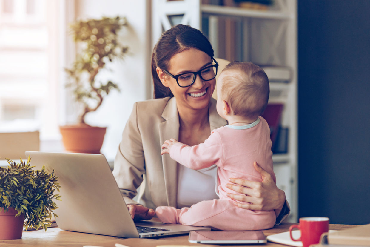 Cheerful young beautiful businesswoman looking at her baby girl with smile while sitting at her working place Cheerful young beautiful businesswoman looking at her baby girl with smile while sitting at her working place