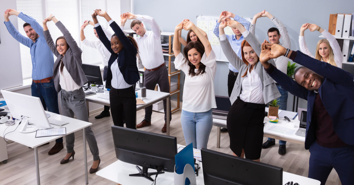A group of office workers stand and stretch at their desks. A group of office workers stand and stretch at their desks.