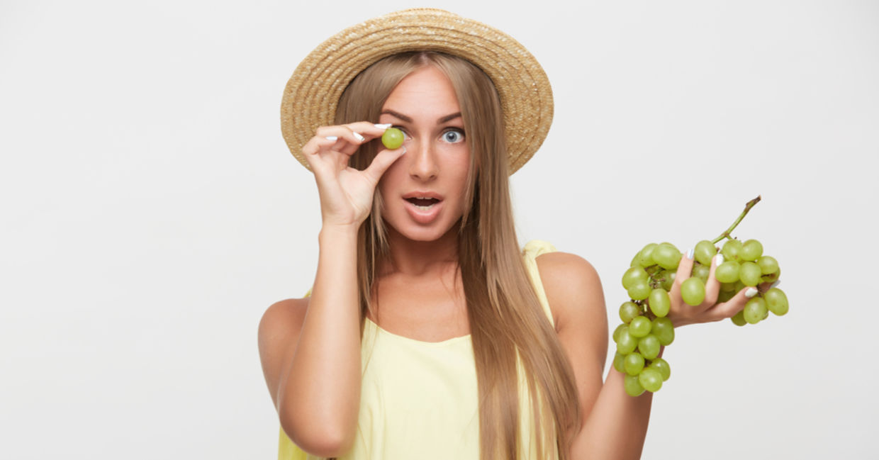 A young woman holds a green grape in front of her eye. A young woman holds a green grape in front of her eye.